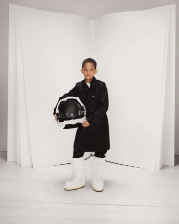 Young boy in a black trench coat holds a space helmet and wears white moon boots in a bright studio with a blank white background.