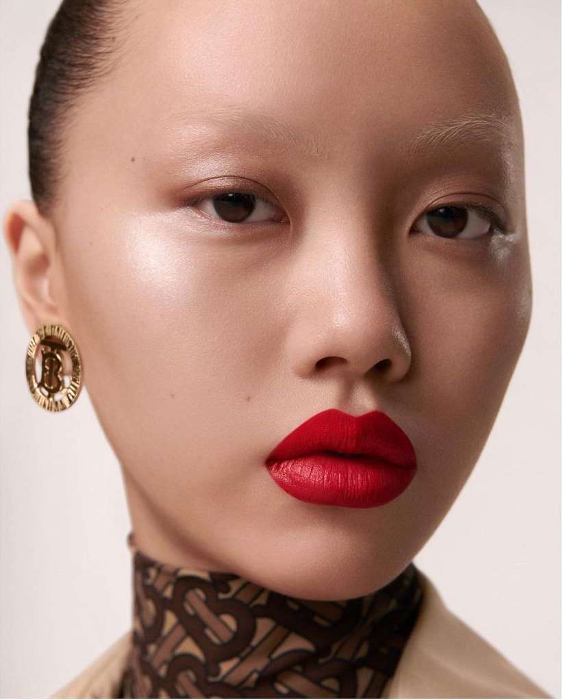 Close-up portrait of an Asian woman with red lipstick and gold earrings against a neutral background, showcasing a beauty advertisement.