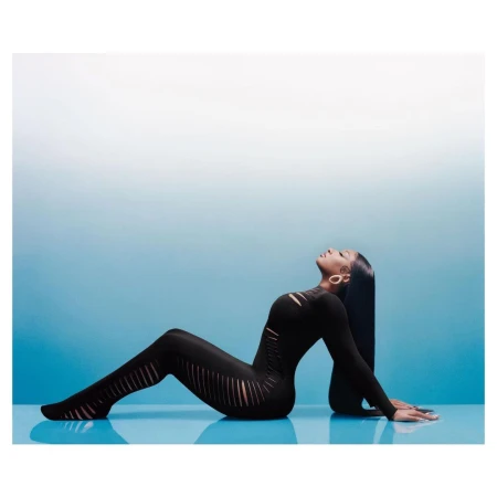 Black woman with long dark hair wearing a black cutout jumpsuit posing against a gradient blue and white backdrop.
