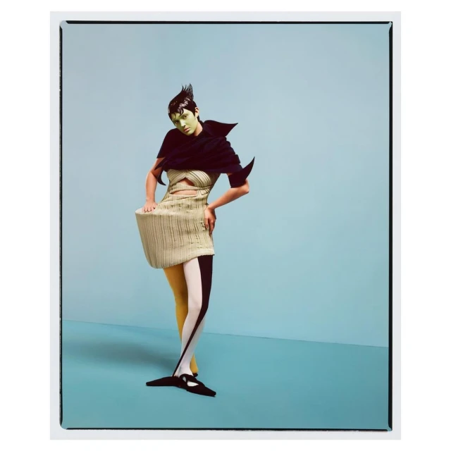 Woman poses in an abstract black top and tan striped dress with patterned stockings against a light blue background in a studio setting.