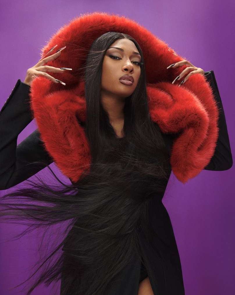 Portrait of a Black woman with long black hair and a fur-trimmed black coat against a purple backdrop in a studio setting.