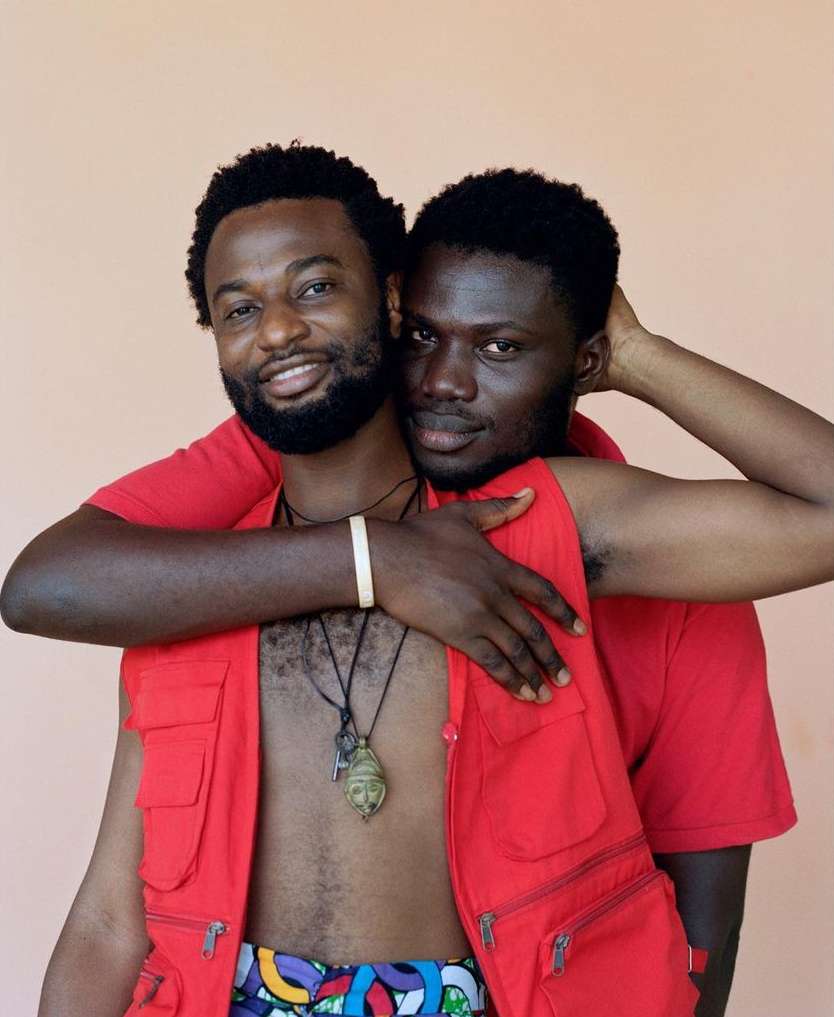 Close-up of two adult men embracing in a photo studio. The man in front wears a red vest; both are smiling against a peach backdrop.