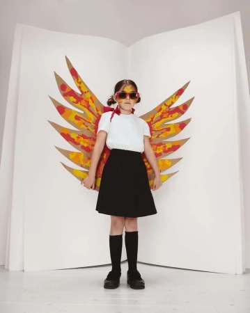 Young girl in white shirt, black skirt, and homemade cardboard wings posing in front of a white backdrop, smiling and wearing sunglasses.