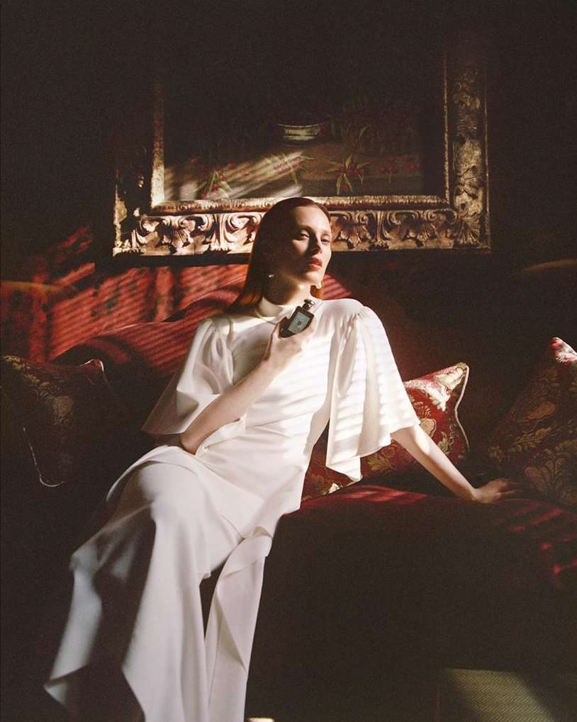 Elegant woman in a white dress sits on a red couch, holding a dark perfume bottle in a moody indoor setting with soft, diffused lighting.