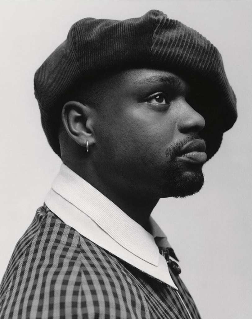 Monochrome portrait of a Black adult male in profile, wearing a cap and checkered jacket, gazing upward against a neutral backdrop.