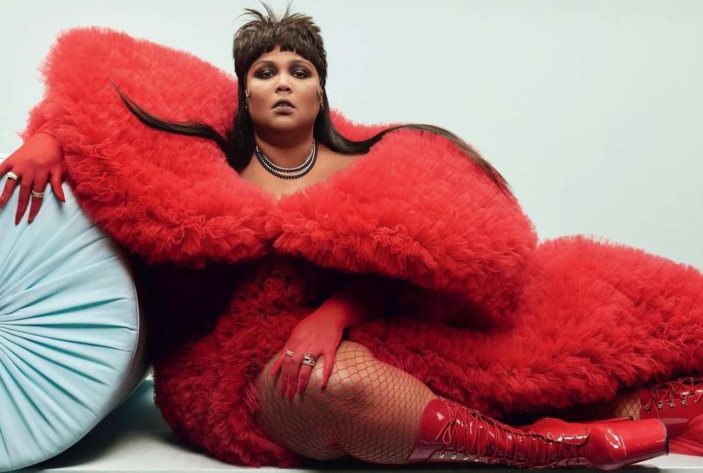 Studio shot of Lizzo reclining on a light blue pillow, wearing red fishnets, red boots, a red textured robe, and jewelry.