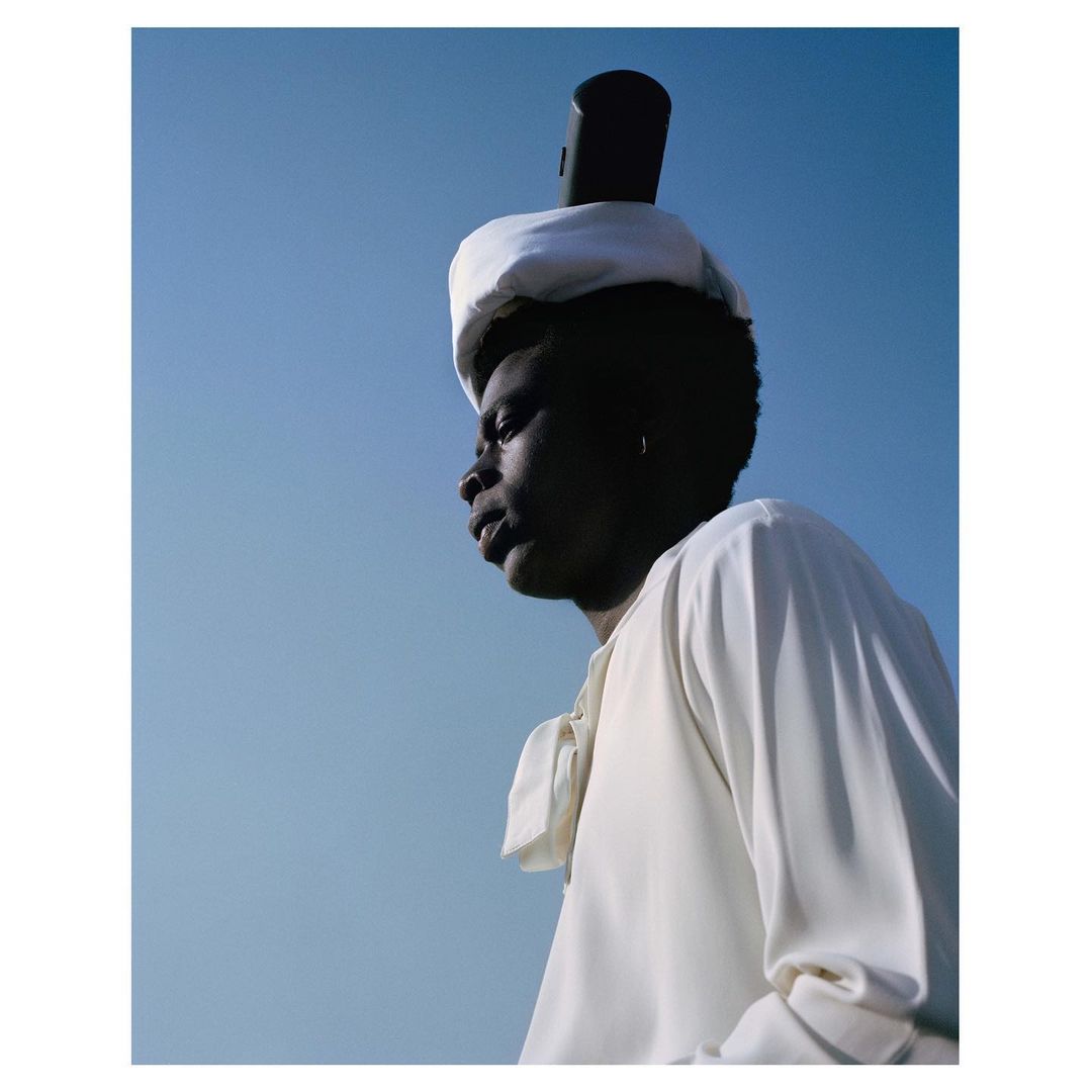 Portrait of a black person in a white outfit, wearing a white hat with a black object balanced on top, against a clear blue sky.