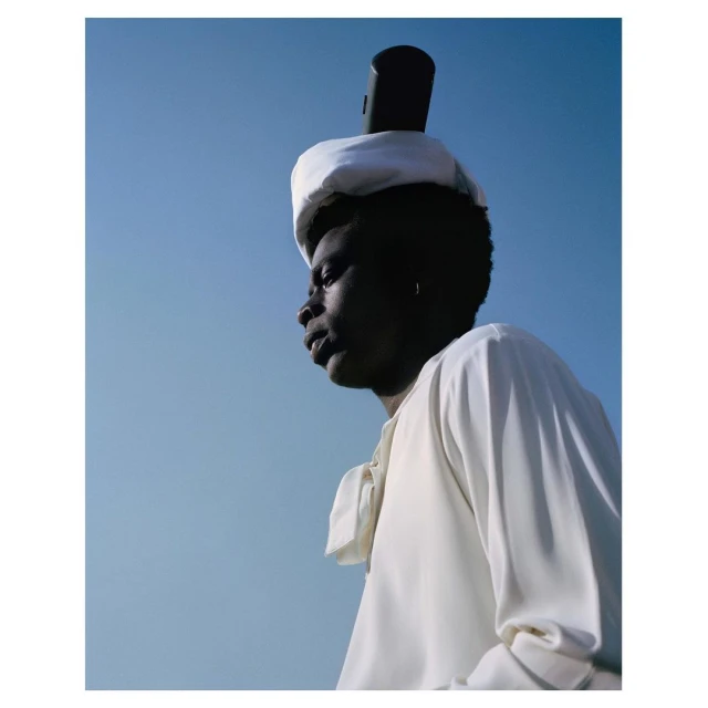Portrait of a black person in a white outfit, wearing a white hat with a black object balanced on top, against a clear blue sky.