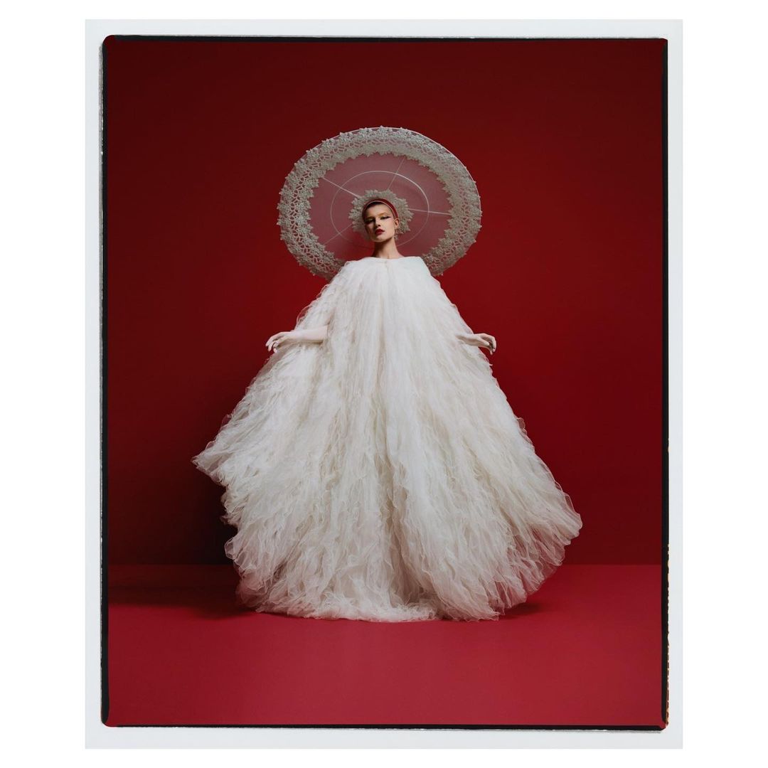 Fashion shot: a model in a white, textured, oversized gown and a white parasol stands in a photo studio against a red backdrop.