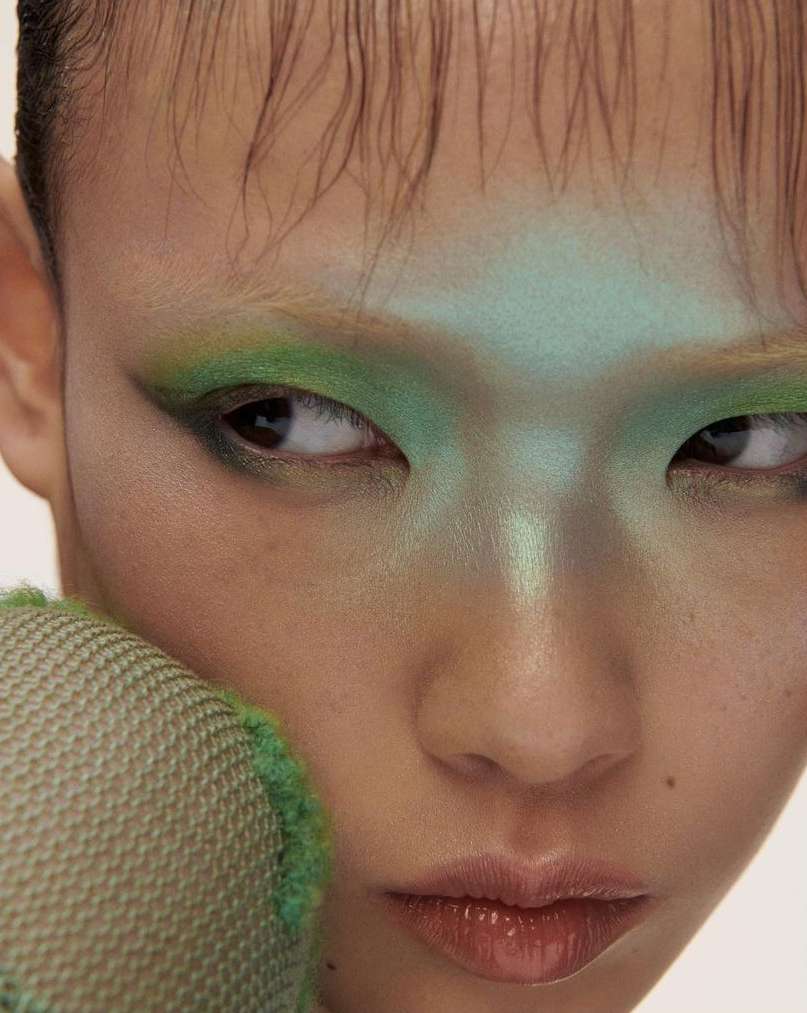 Close-up portrait of a young person with artistic green eyeshadow and light green makeup on the forehead, showcasing beauty and style.