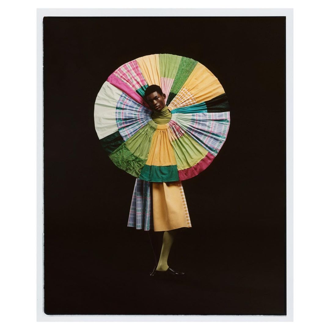 Fashionable Black model poses in a colorful, patterned dress with matching legwear against a dark backdrop in a photo studio setting.