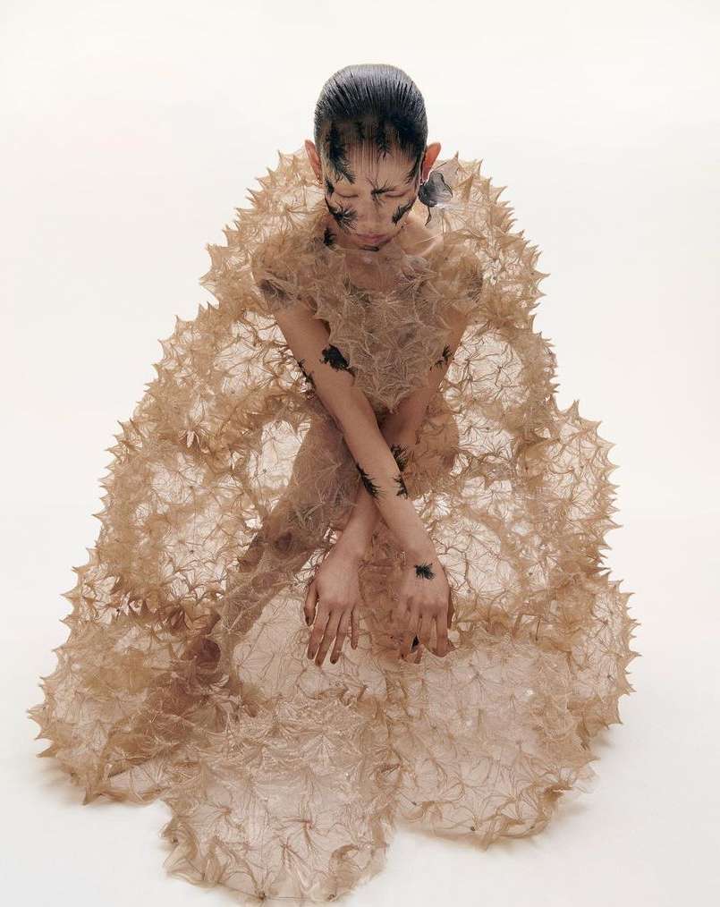 Asian female model in avant-garde fashion kneeling in a light photo studio with creative eye and arm makeup and unique beige spiky gown.