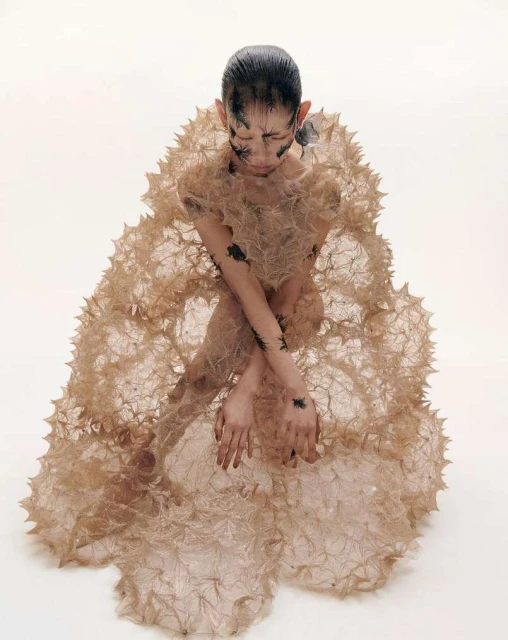 Asian female model in avant-garde fashion kneeling in a light photo studio with creative eye and arm makeup and unique beige spiky gown.