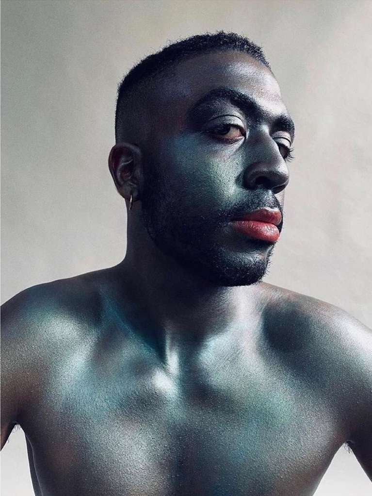 Striking portrait of a Black adult male with face and body makeup, wearing a gold hoop earring against a simple, light background.