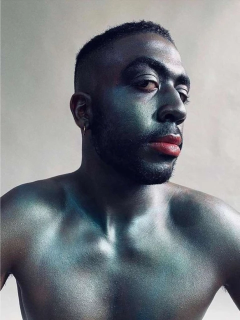 Striking portrait of a Black adult male with face and body makeup, wearing a gold hoop earring against a simple, light background.