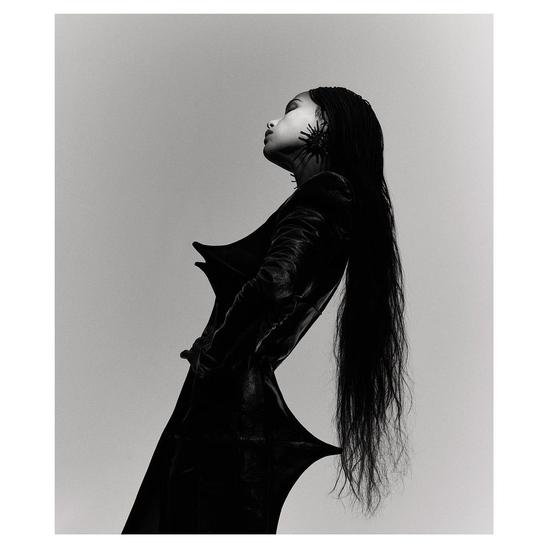 Monochrome fashion shot of a woman with long black hair wearing a black dress and statement earrings against a grey background.
