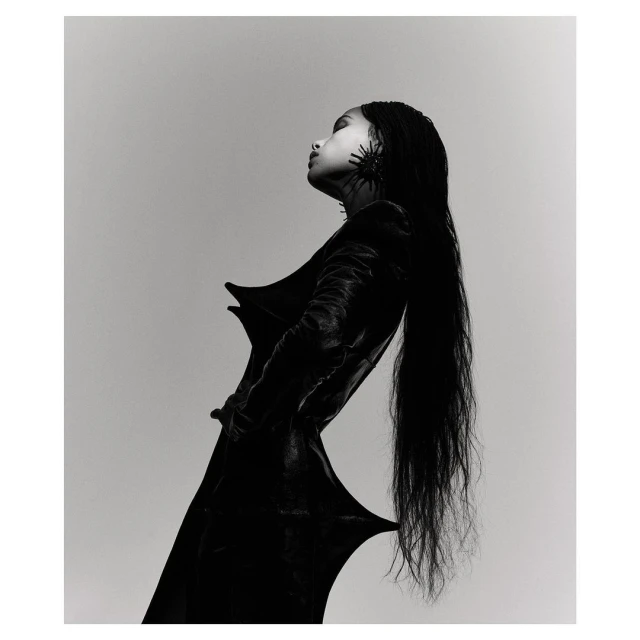 Monochrome fashion shot of a woman with long black hair wearing a black dress and statement earrings against a grey background.