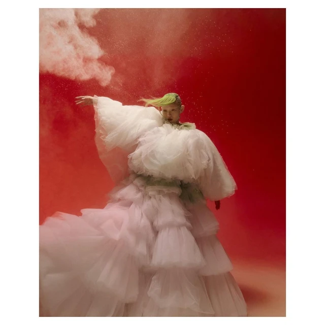 Woman in voluminous ruffled white dress posing against a vibrant red background with a wispy cloud of pink powder.