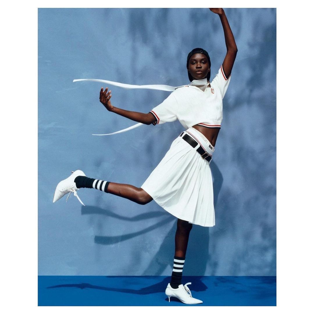 Black female model wearing a white polo shirt and pleated skirt dancing against a blue backdrop
