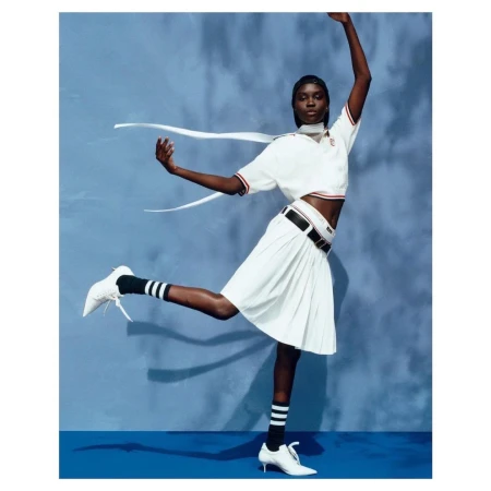 Black female model wearing a white polo shirt and pleated skirt dancing against a blue backdrop
