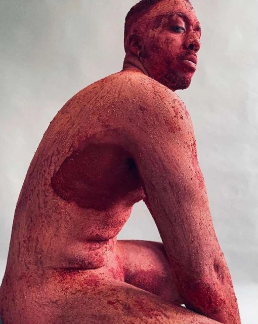 Nude adult male, coated in red sand, sits in a studio with an expressive pose, looking back over his shoulder towards the camera.