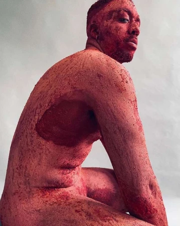 Nude adult male, coated in red sand, sits in a studio with an expressive pose, looking back over his shoulder towards the camera.