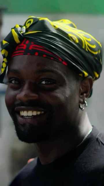 Close-up shot of a smiling African American man wearing a yellow and black patterned headwrap, earring and black t-shirt.
