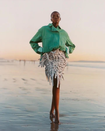 Model standing on a sandy beach at sunset wearing a green jacket and silver fringed skirt, captured by Campbell Addy.