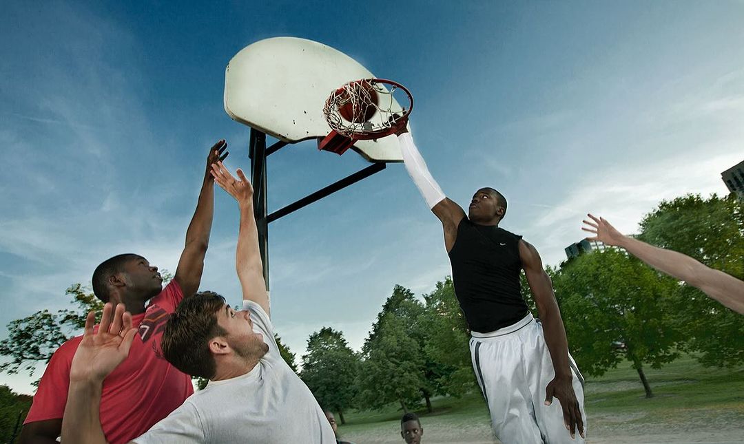 Four adult male basketball players in action during a game in a grassy park with trees against a blue sky backdrop with a basketball hoop.