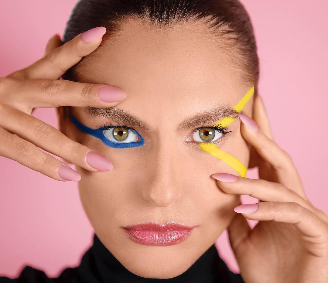 Close-up studio shot of a model with artistic blue and yellow makeup around her eyes, complemented by pink manicured nails against pink backdrop.