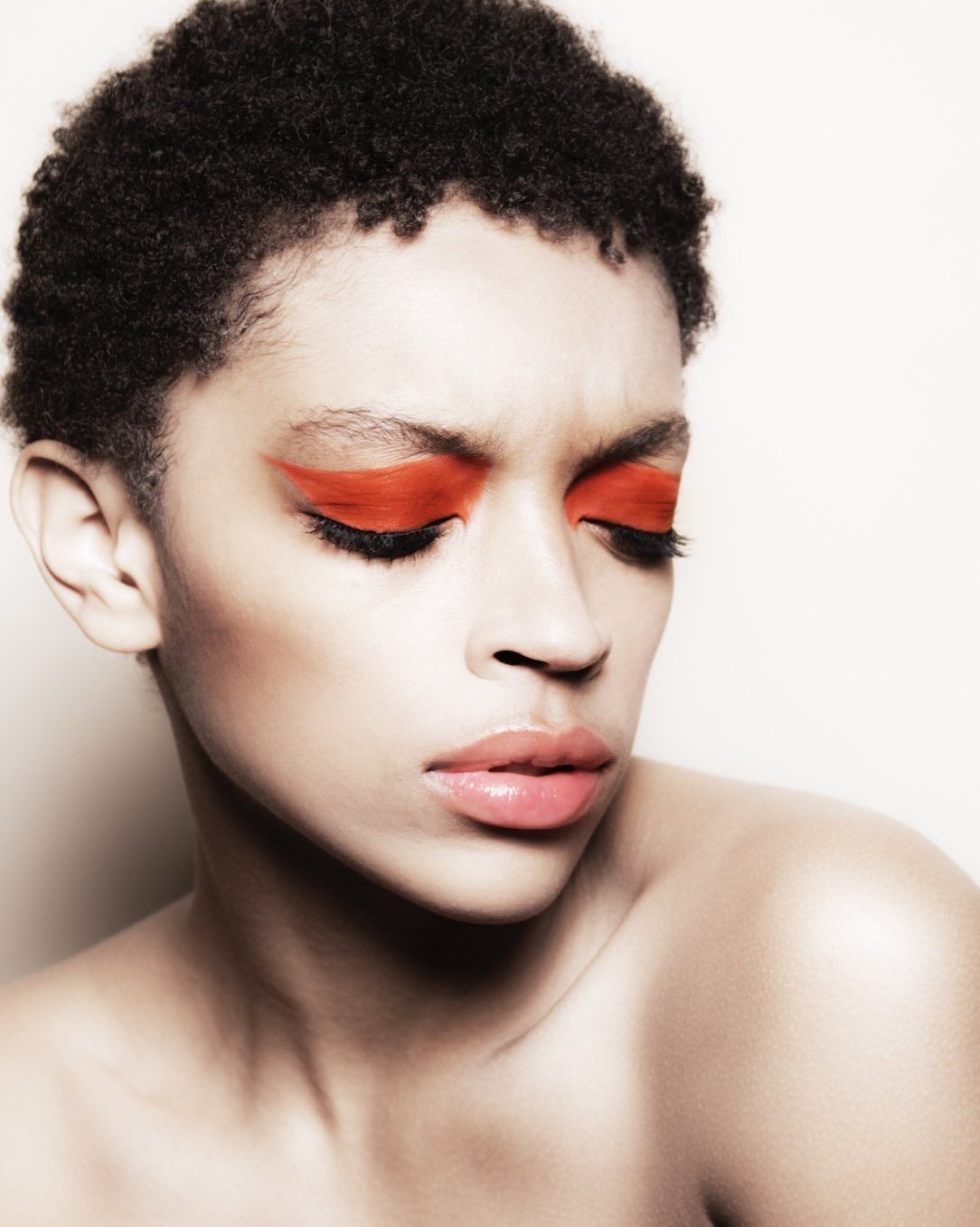 Close-up portrait of a Black woman with orange geometric eye makeup and glossy lips against a light background in beauty shot.
