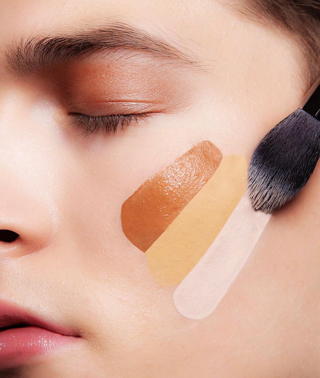 Close-up of a person's face having foundation applied with a brush, showcasing different shades on the cheek.