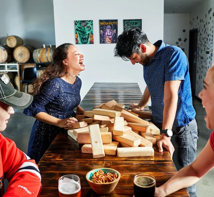 Four young adults laugh and play a stacking-blocks game on a wooden table indoors. They are dressed casually and seem to be having fun.