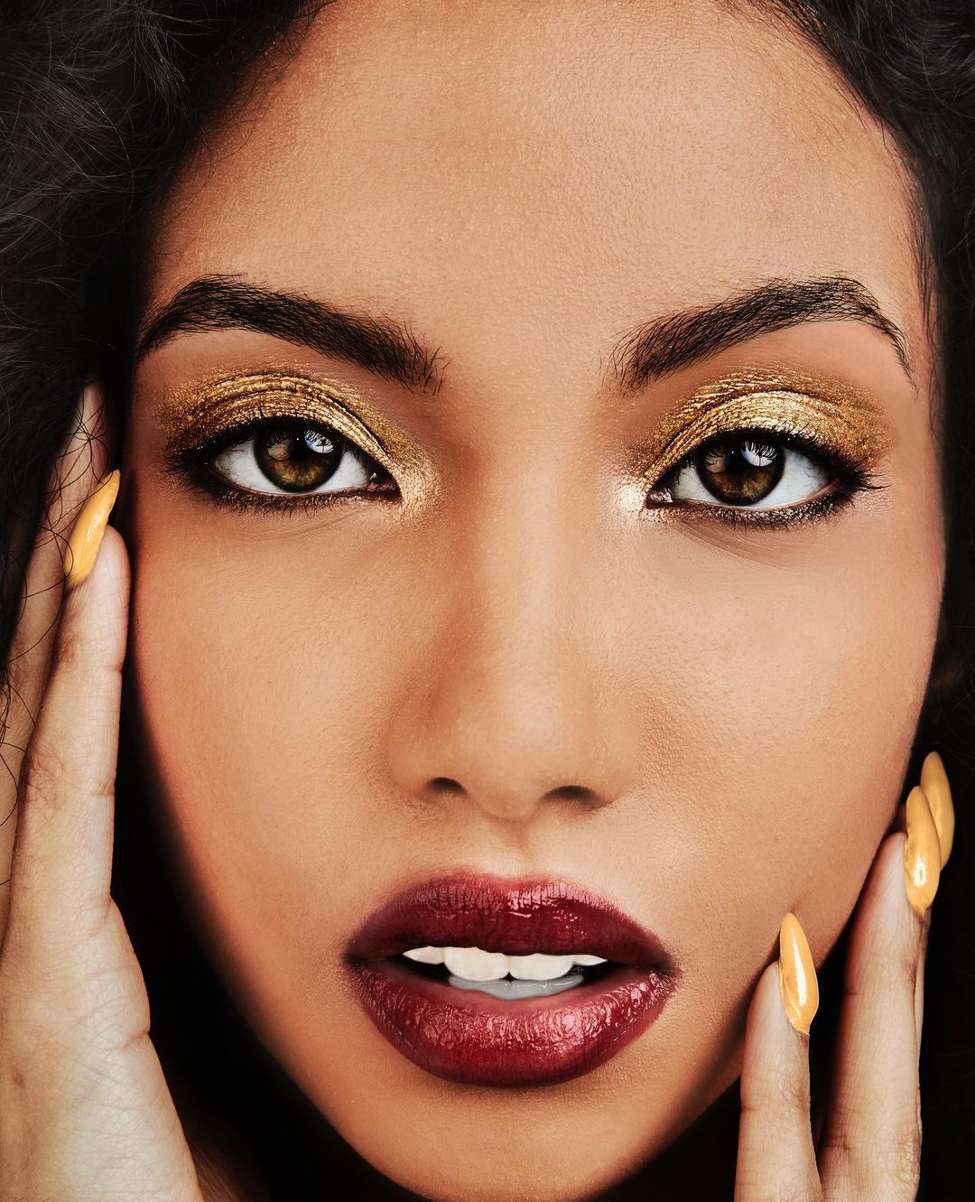 Close-up portrait of a woman with gold eyeshadow, dark red lipstick, and yellow nails, touching her face with both hands indoors.