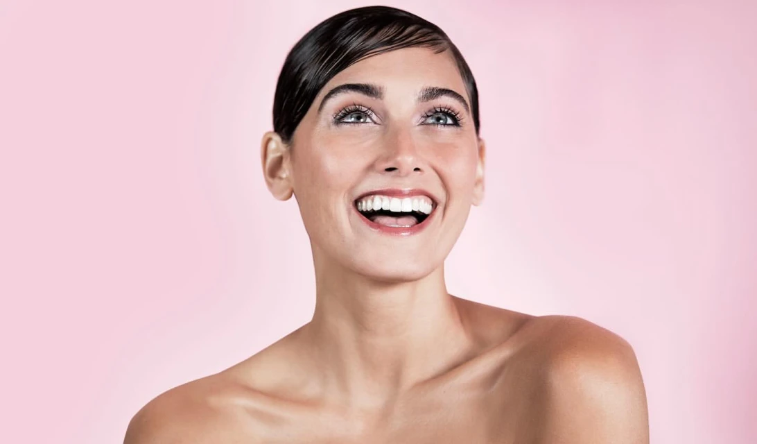 Close-up beauty shot of a smiling woman with wet-look hair against a pink background, looking up, showcasing beauty and skincare.