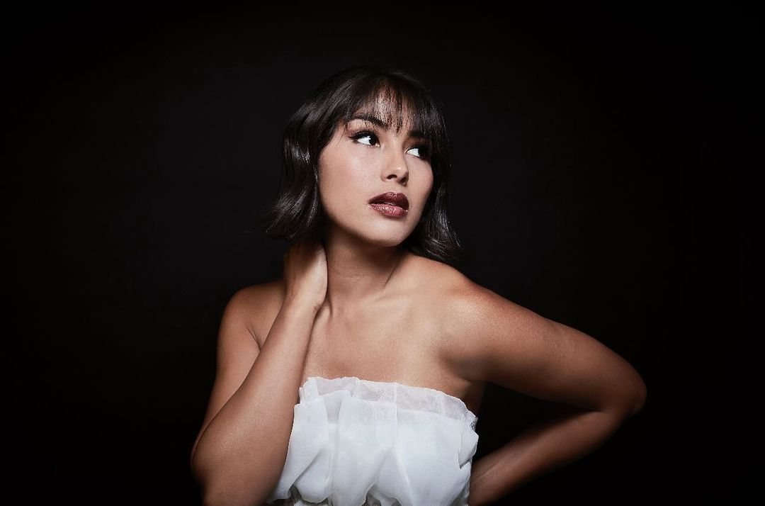 Portrait of a brunette woman with bangs looking off-camera, wearing a white ruffled top against a dark background in a photo studio.