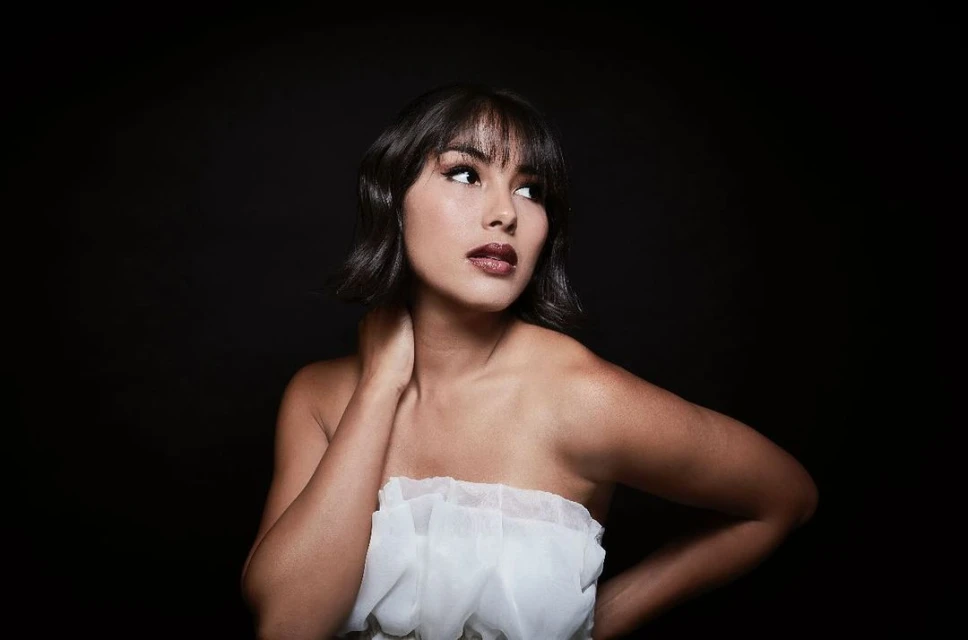 Portrait of a brunette woman with bangs looking off-camera, wearing a white ruffled top against a dark background in a photo studio.