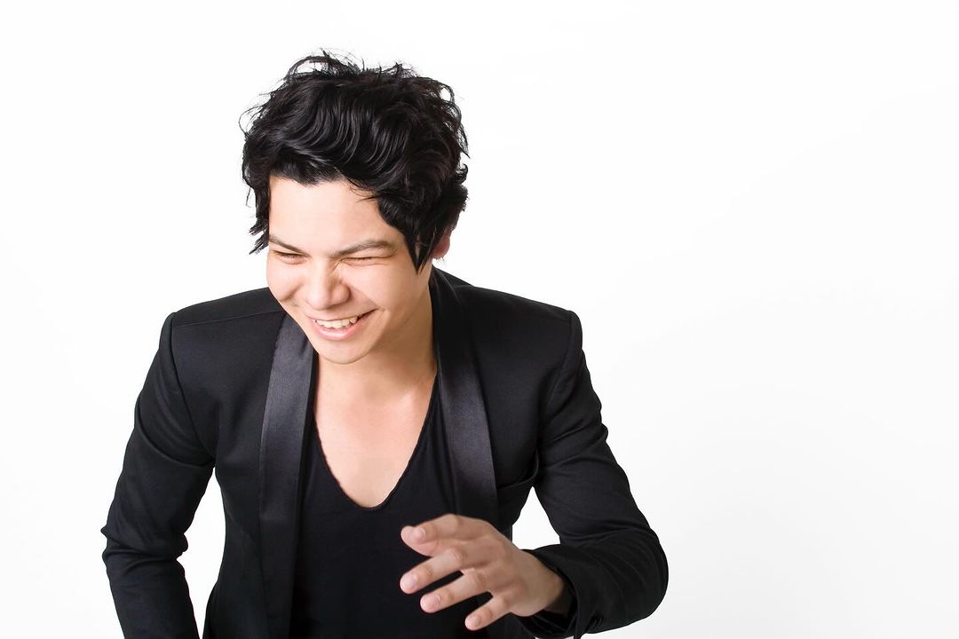 Portrait of a smiling adult male with dark hair, wearing a black shirt and jacket, against a bright white background in a photo studio.
