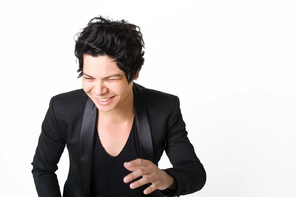 Portrait of a smiling adult male with dark hair, wearing a black shirt and jacket, against a bright white background in a photo studio.