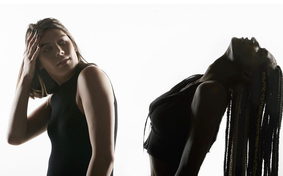 Two adult women in a studio pose against a bright white background; one with braids arched back, one with hand on head.
