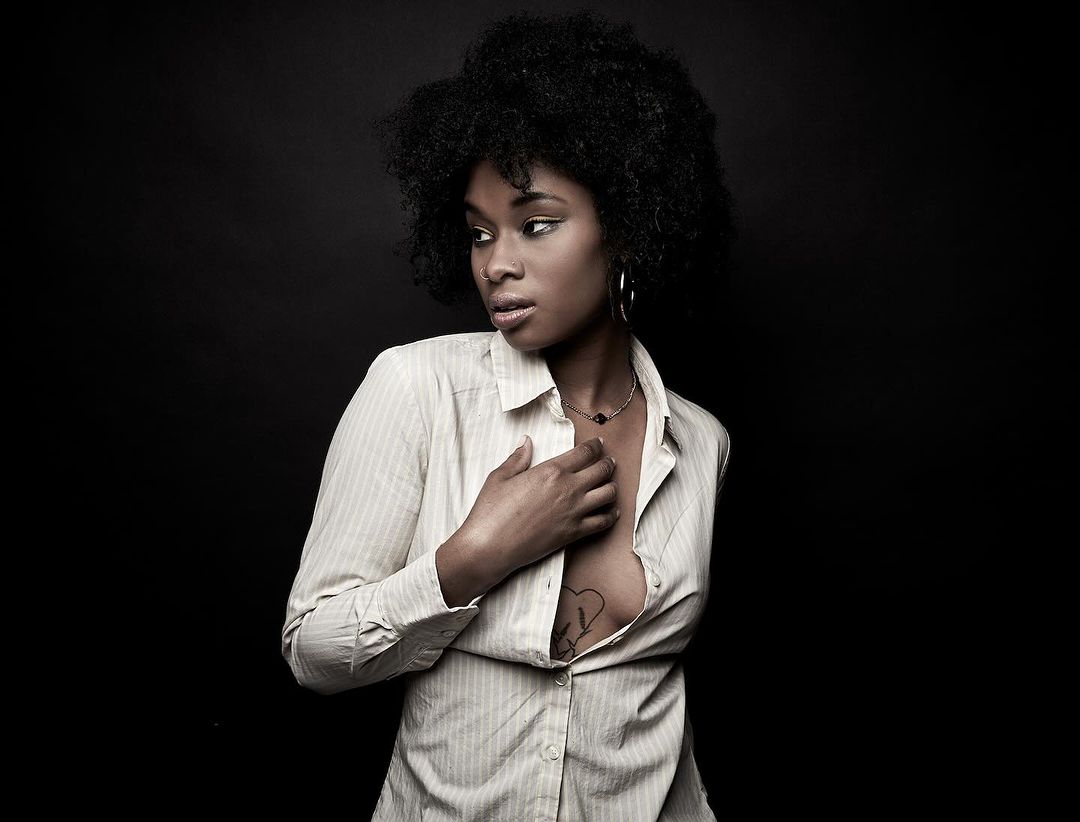 Portrait of African American woman with afro hairstyle in unbuttoned striped shirt, wearing jewelry, against a dark studio backdrop.