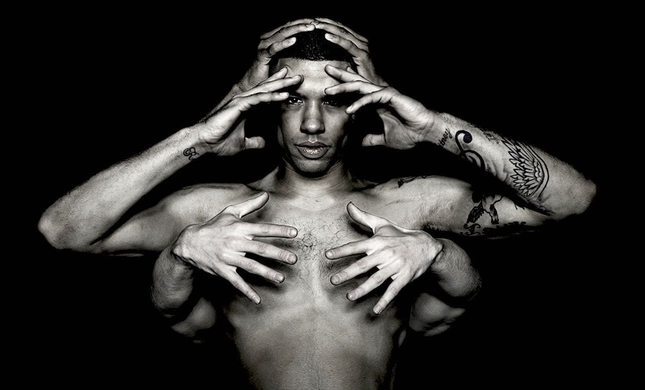 Striking black and white portrait of a shirtless adult man with multiple arms and tattooed forearms against a dark black background.