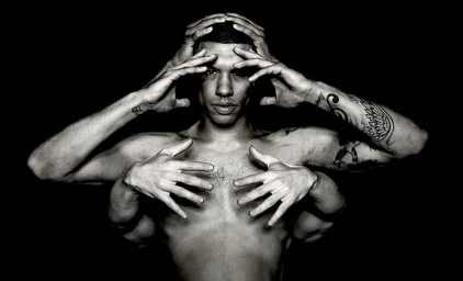 Black and white portrait of a man with multiple arms posed around his head and chest against a dark background.