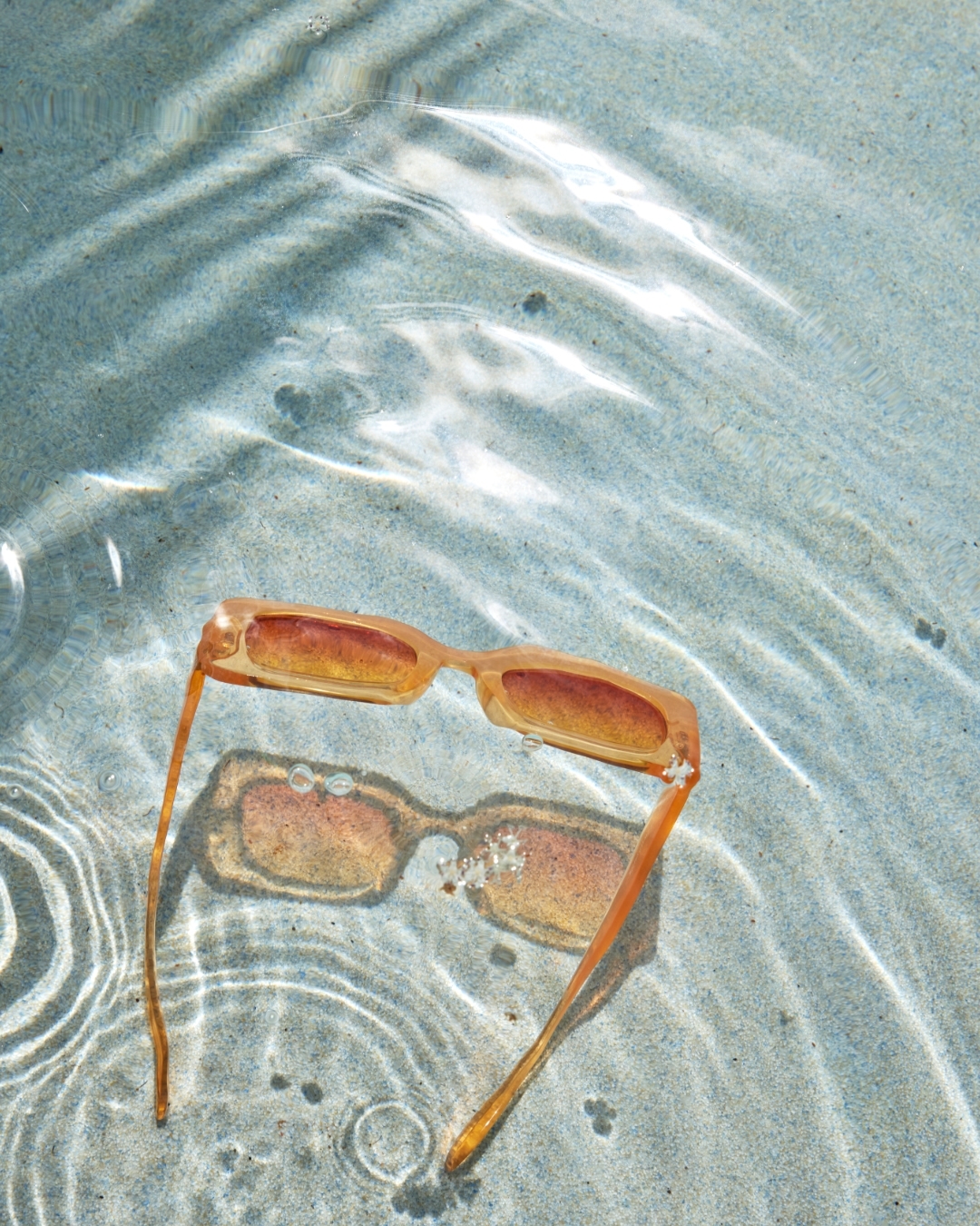Peach-colored sunglasses rest on a light blue textured surface with water ripples, creating a cool, summery vibe in natural light.