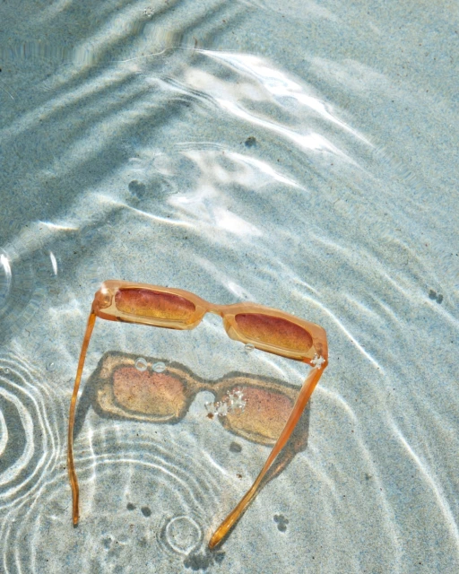 Peach-colored sunglasses rest on a light blue textured surface with water ripples, creating a cool, summery vibe in natural light.