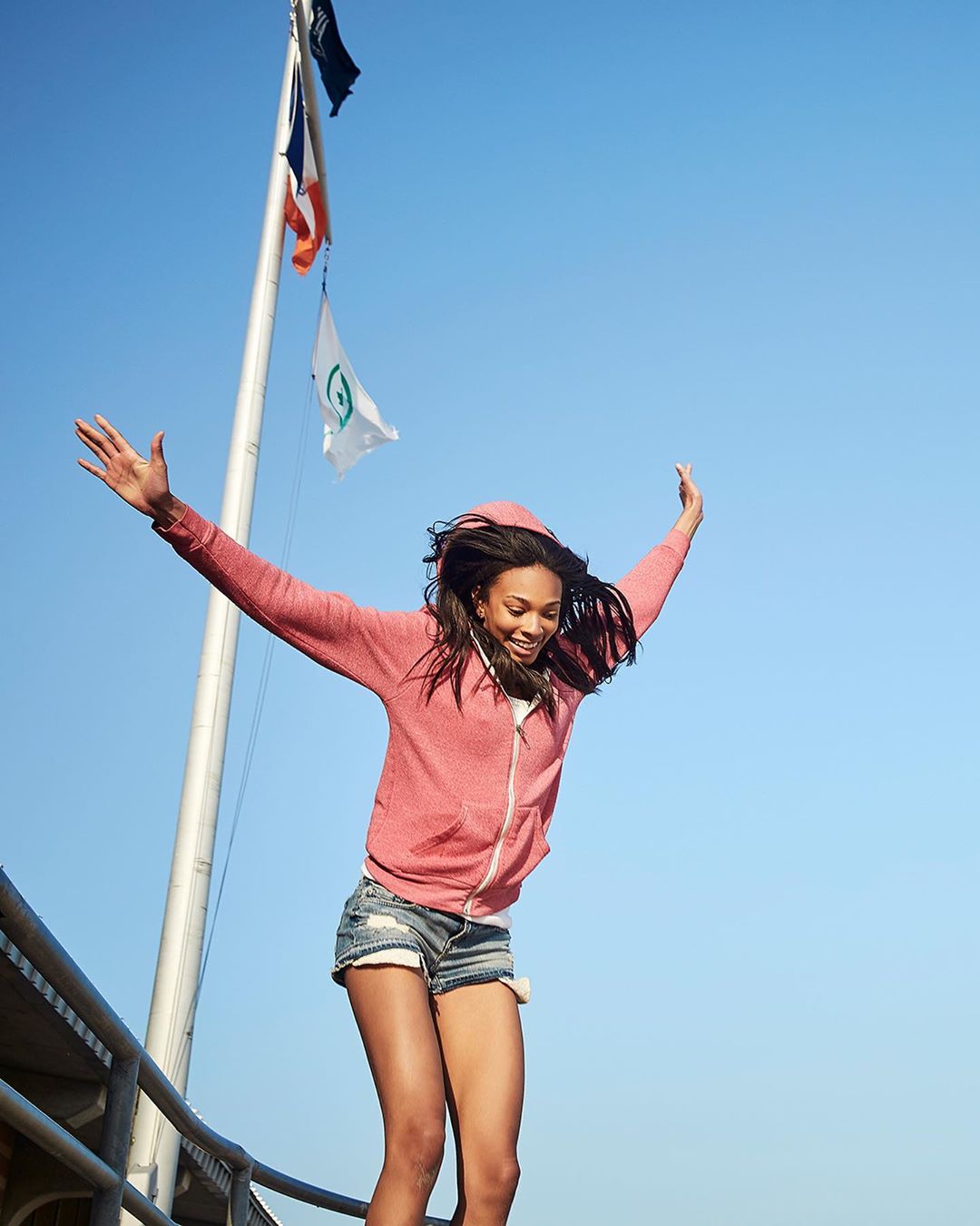 Young woman in shorts and a hoodie jumps in the air with arms outstretched against a clear blue sky, flags and metal railing visible.