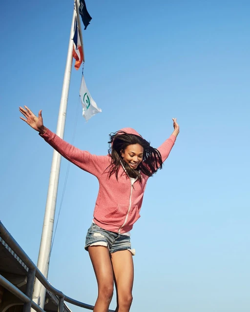 Young woman in shorts and a hoodie jumps in the air with arms outstretched against a clear blue sky, flags and metal railing visible.