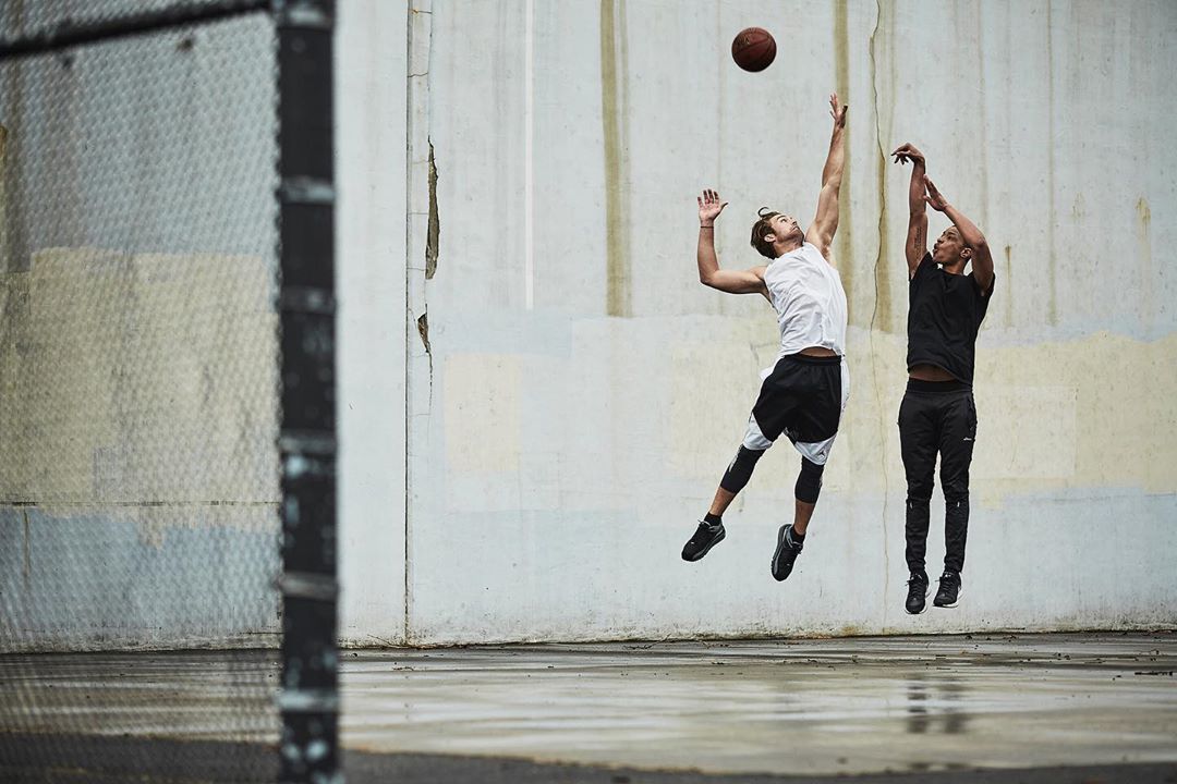 Two adult male basketball players jumping to catch the basketball outside with a concrete background and a chain-link fence on the left.
