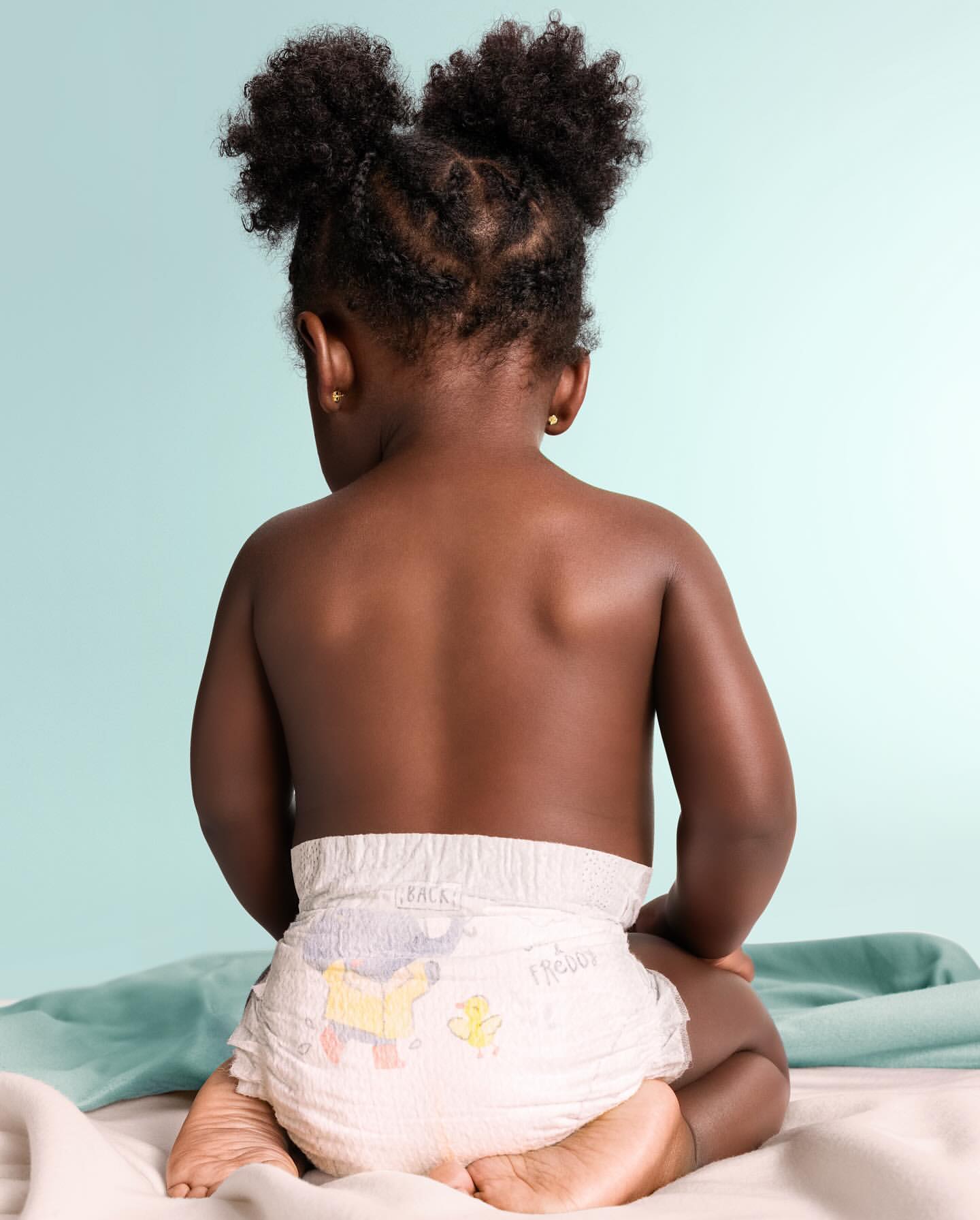 Adorable African-American toddler with afro puffs, seated with back to camera, wearing a diaper, on a soft blanket against an aqua background.