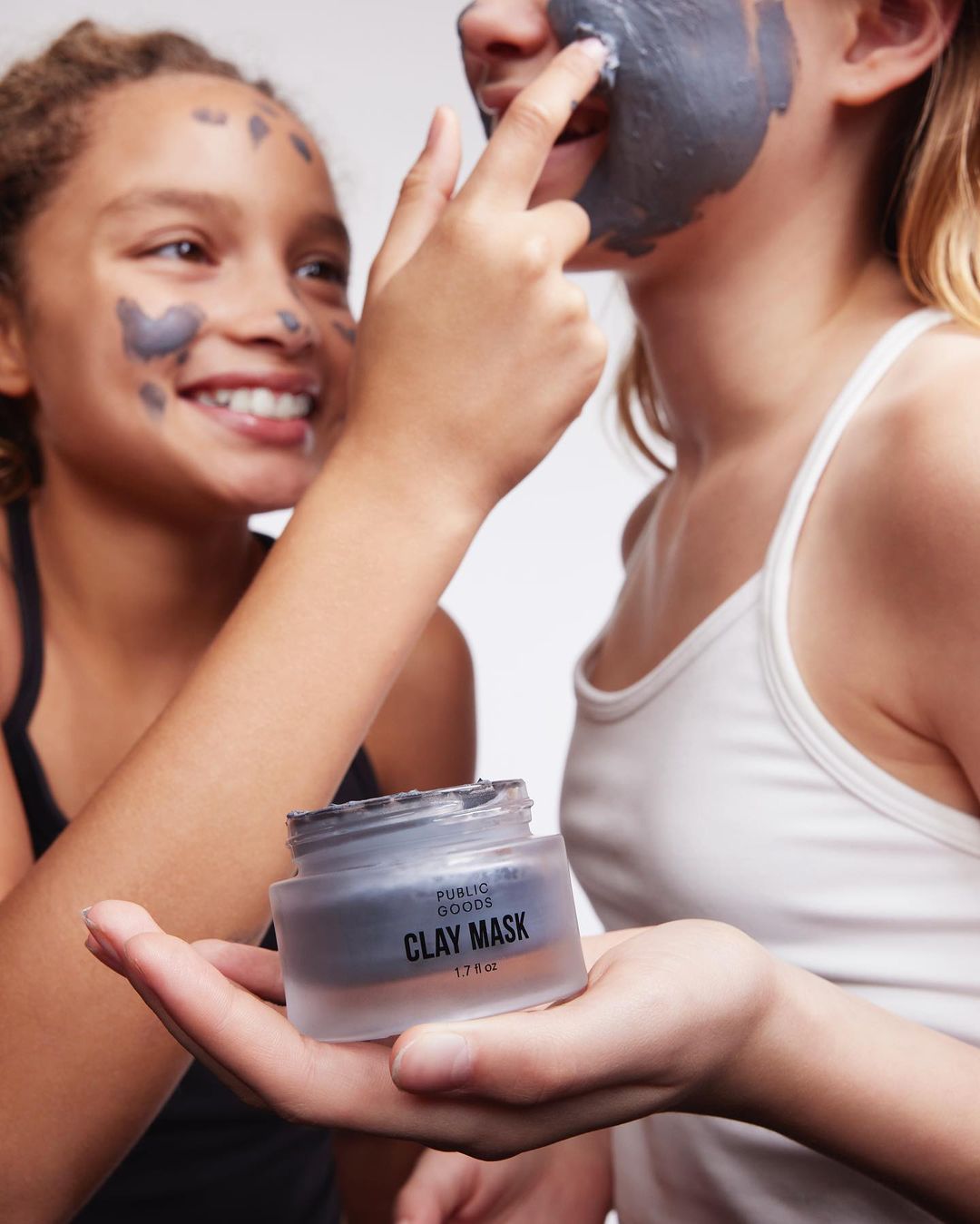 Two girls applying a grey clay mask to their faces while indoors, holding the jar, with a bright white background.